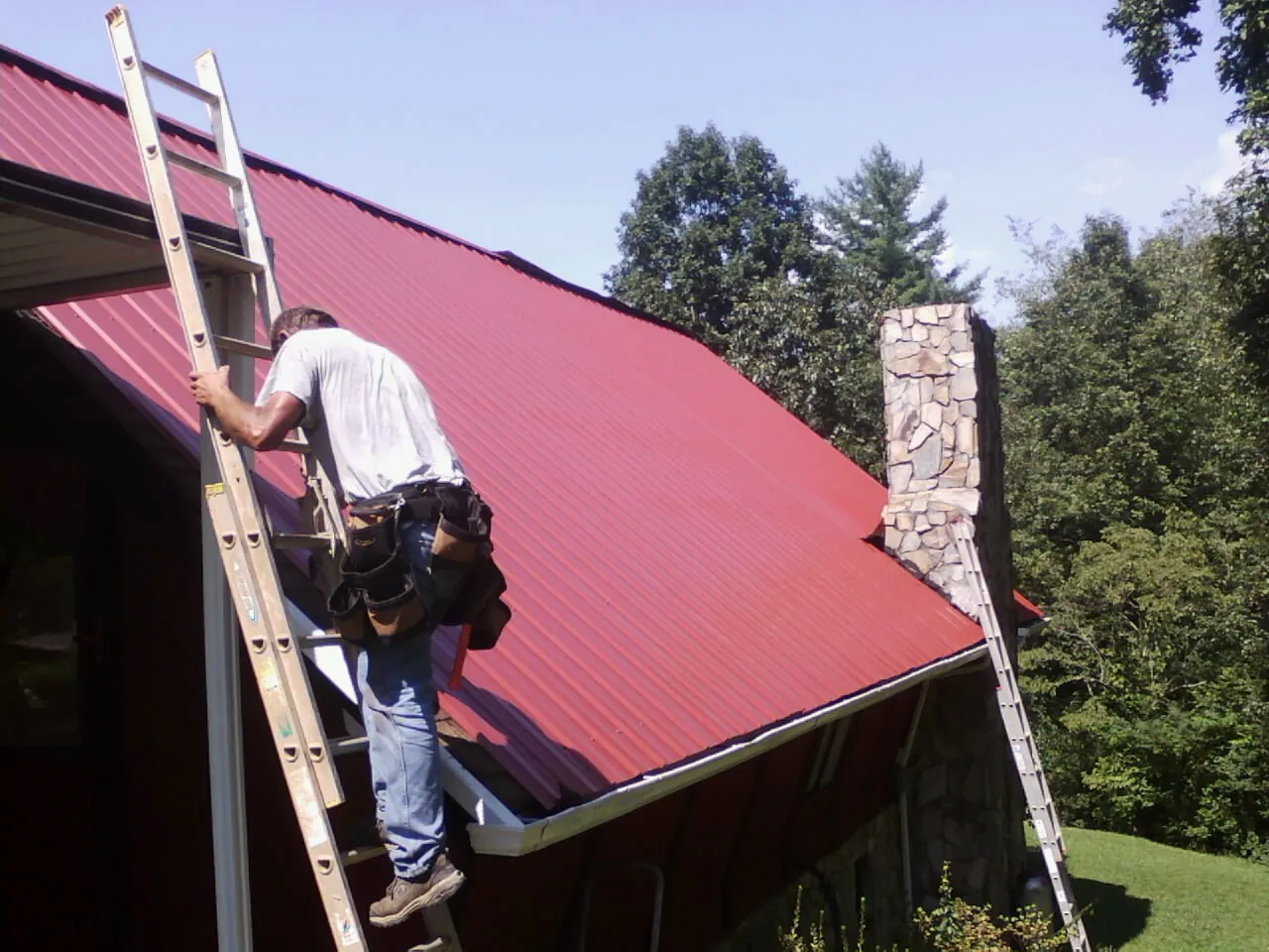 Roofer on a ladder beside a red metal roof and stone chimney — Soffit & Fascia Repair by Roofing Brothers in New Baltimore